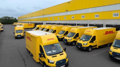 A line of yellow DHL delivery vans parked along the dock of the yellow DHL warehouse.