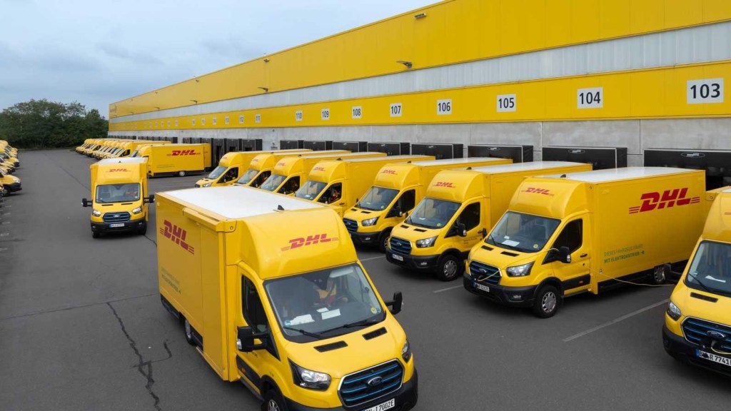 A line of yellow DHL delivery vans parked along the dock of the yellow DHL warehouse.