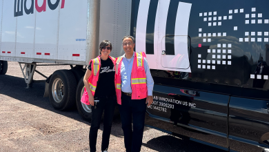 Two people wearing bright pink safety vests and smiling while posing in front of Waabi-branded semi-truck trailers on a sunny gravel lot.