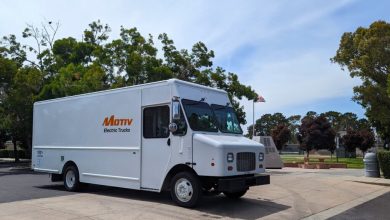 A Motiv Electric Trucks white medium-duty electric delivery truck parked outdoors under a blue sky, with trees and an American flag in the background, illustrating the company's role in the merger with Workhorse Group.