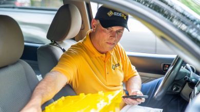 Inside car, view of delivery driver wearing gold shirt with black hat that reads SDS Rx