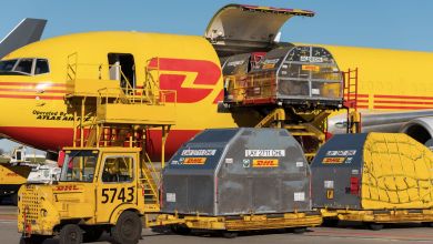 A bright yellow DHL cargo jet with side cargo door open as containers are loaded with hydraulic lifts.