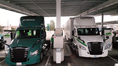 Two electric trucks, one branded with "Nevoya" and the other with "Penske Electric Vehicle," are parked under a covered charging station at a Greenlane facility. Charging equipment is connected to both trucks, and personnel in green uniforms are attending to the chargers. The scene includes a modern charging infrastructure setup with a clear sky and some distant buildings in the background.