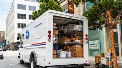 Rear view of a USPS delivery van with rear door open, packages inside, parked on city street next to a UPS Store.