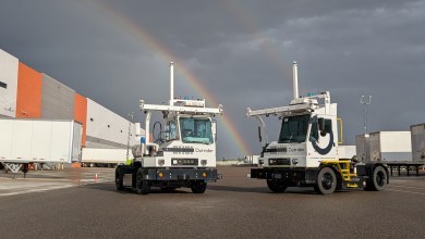 Two Outrider autonomous yard trucks in a logistics facility under a double rainbow, showcasing secure AI-powered yard operations technology following SOC 2 Type 2 attestation.