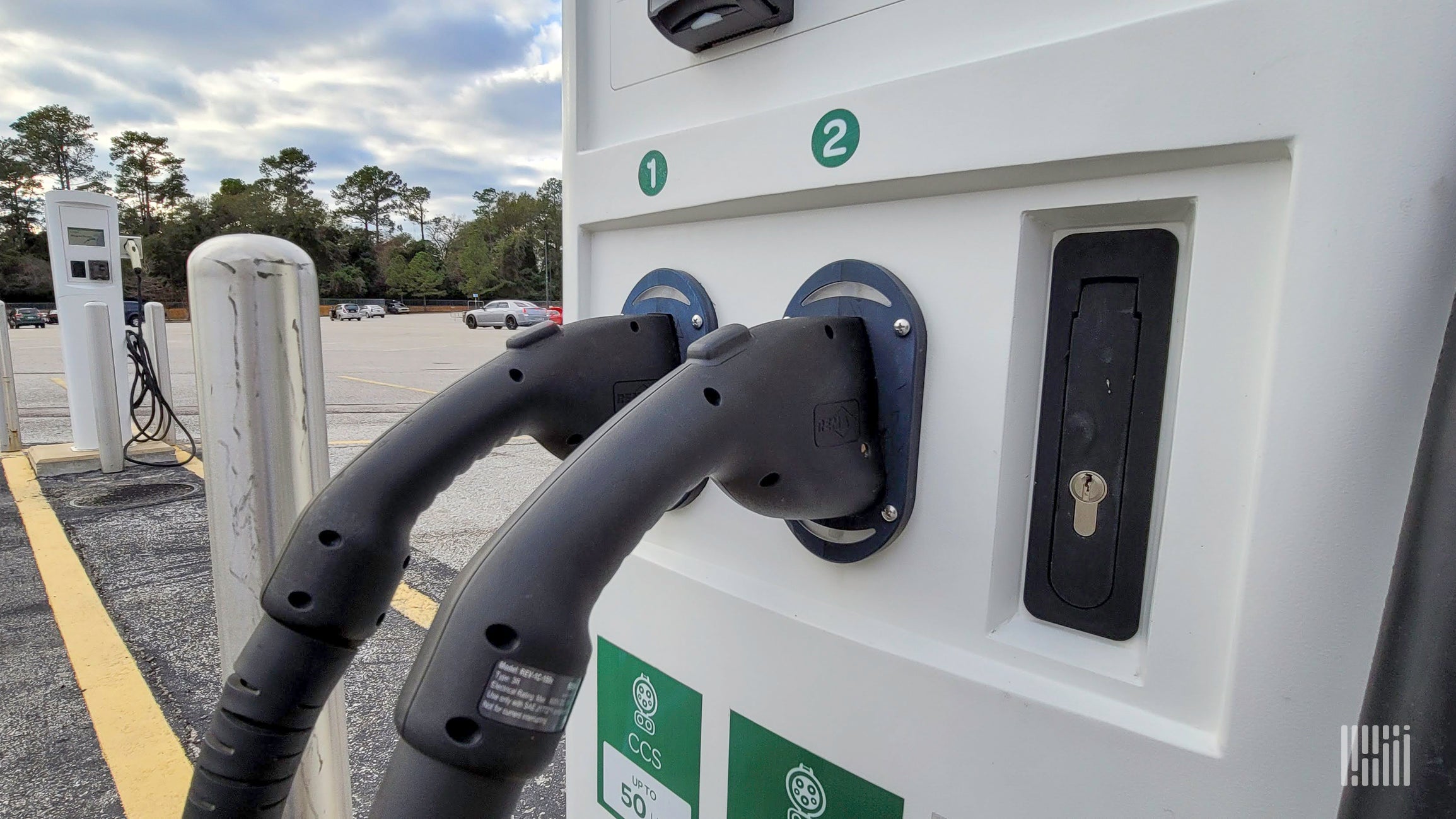 A close-up view of a white electric vehicle charging station with two black CCS plugs labeled 1 and 2, rated at 50 kW, in an outdoor parking lot with cars and trees in the background.