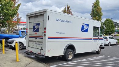 Side-rear view of a USPS van in a strip mall parking spot.