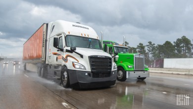 A white Schneider tractor pulling an orange intermodal container on a rainy highway next to a neon green tractor