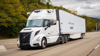 A white Volvo VNL Autonomous semi-truck, branded with Waabi and Volvo Autonomous Solutions logos, drives on a highway amid autumn foliage, showcasing advanced self-driving technology.