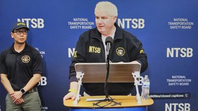 An NTSB official stands at a podium to talk with reporters with NTSB logo in background.