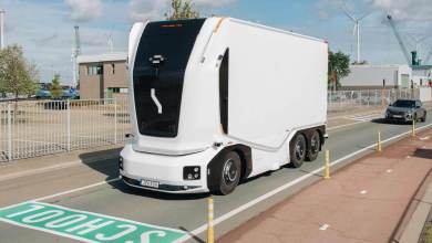 A futuristic white cabless autonomous electric truck from Einride drives on a public road in an industrial area, with wind turbines, cranes, and buildings in the background, under a partly cloudy sky. A small gray car follows behind, and yellow cones line the lane near a green "SCHOOL" marking on the pavement.