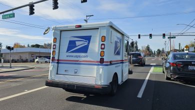 Rear view of a USPS van driving through an intersection with traffic lights.