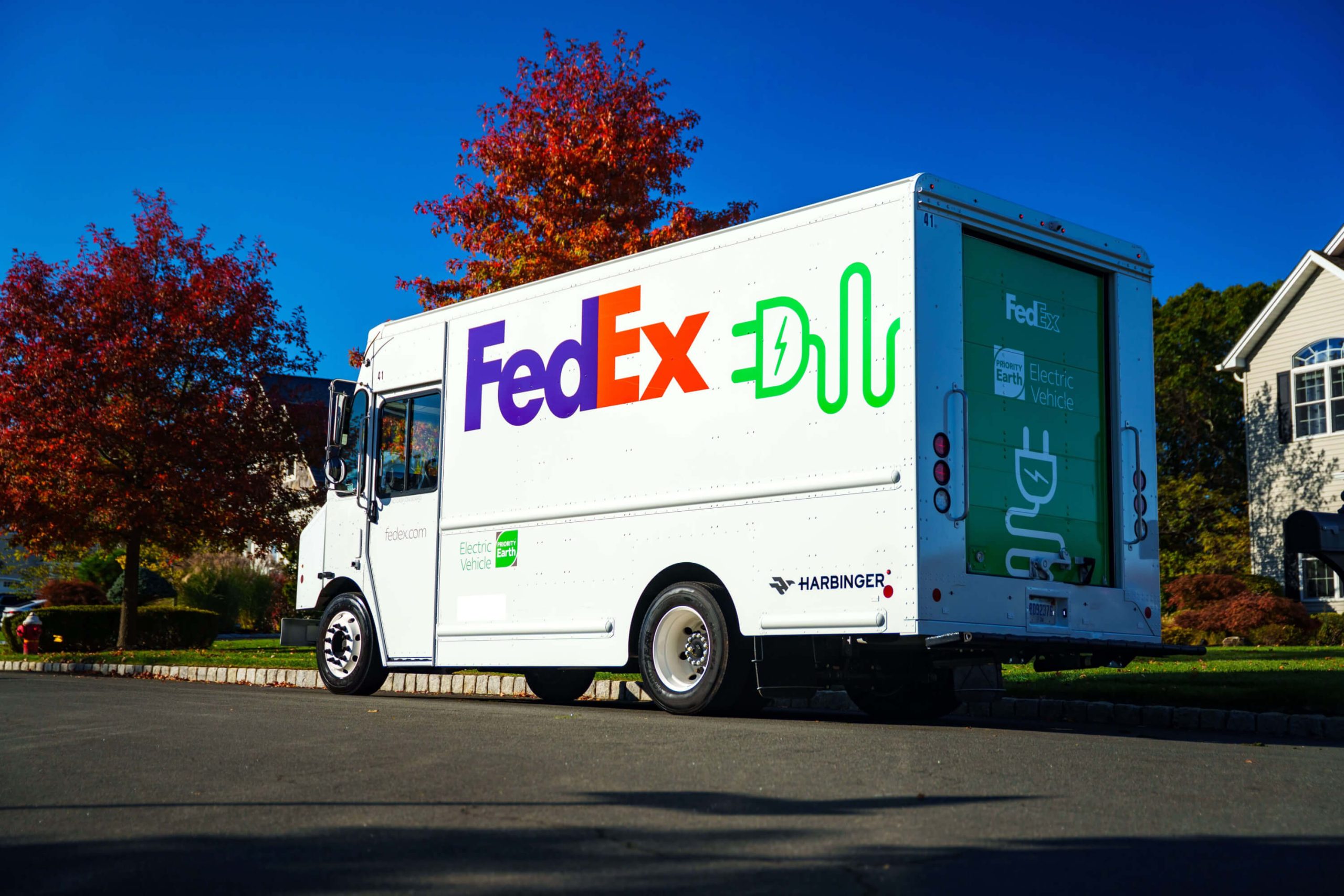 A white FedEx electric delivery truck manufactured by Harbinger, branded with green electric plug icons and "FedEx Earth Electric Vehicle" lettering, parked on a suburban street with autumn foliage and blue sky.