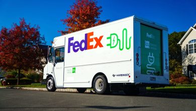A white FedEx electric delivery truck manufactured by Harbinger, branded with green electric plug icons and "FedEx Earth Electric Vehicle" lettering, parked on a suburban street with autumn foliage and blue sky.