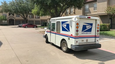 A rear view of a USPS van delivering mail at an apartment complex on a sunny day.