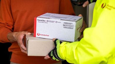 A postal carrier in a yellow pancho delivers an Australia Post parcel to a customer at their door.
