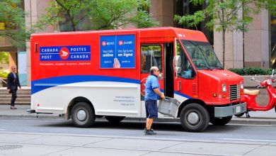 A red Canada Post delivery van with blue and white accents is on a city street as the driver enters the side door.