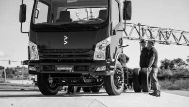Black-and-white photo of a Bollinger Motors electric commercial truck parked roadside, featuring its rugged chassis cab design and company logo, with two men in work attire standing beside the front tire, industrial bridge in the background.