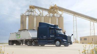 A sleek navy blue Aurora Driver-powered autonomous truck from Detmar Logistics hauls a load of frac sand at a Permian Basin proppant facility, positioned beside towering yellow silos and conveyor belts under a clear blue sky, highlighting innovations in driverless trucking for oil and gas operations.