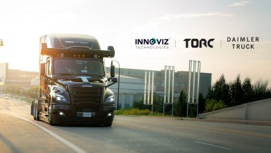 A sleek black Freightliner Cascadia autonomous semi-truck drives along a sunlit urban highway at dawn, featuring Torc Robotics and Daimler Truck branding on the roof, alongside Innoviz Technologies LiDAR sensors on the cab.