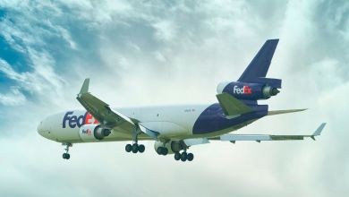 A purple-tailed tri-engine FedEx jet approaches for landing, as seen from rear angle with white cloud wisps in the air.