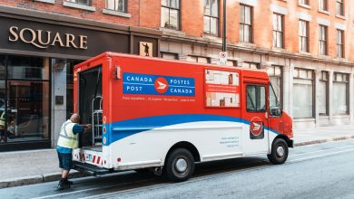 A red-white-and-blue Canada Post truck on a city street, with the driver unloading from the rear.