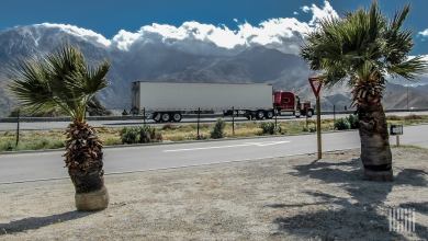 A red semi-truck hauling a white trailer drives along a California highway lined with palm trees, with dramatic mountains in the background under a partly cloudy sky. This image represents the future of heavy-duty autonomous trucks in California as the DMV's revised 2025 regulations open the door to driverless big rigs and self-driving semis on state roads.