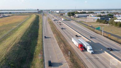 Aerial view of a multi-lane interstate highway with several semi-trucks and passenger vehicles traveling in light traffic under clear skies, representing the operational environment for autonomous trucking technology and product liability risks in the freight industry.