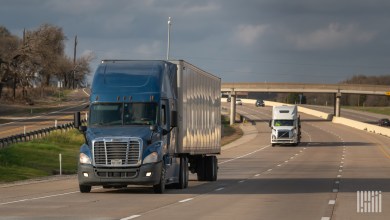 a blue tractor pulling a trailer on a highway with a white tractor trailing in the distance