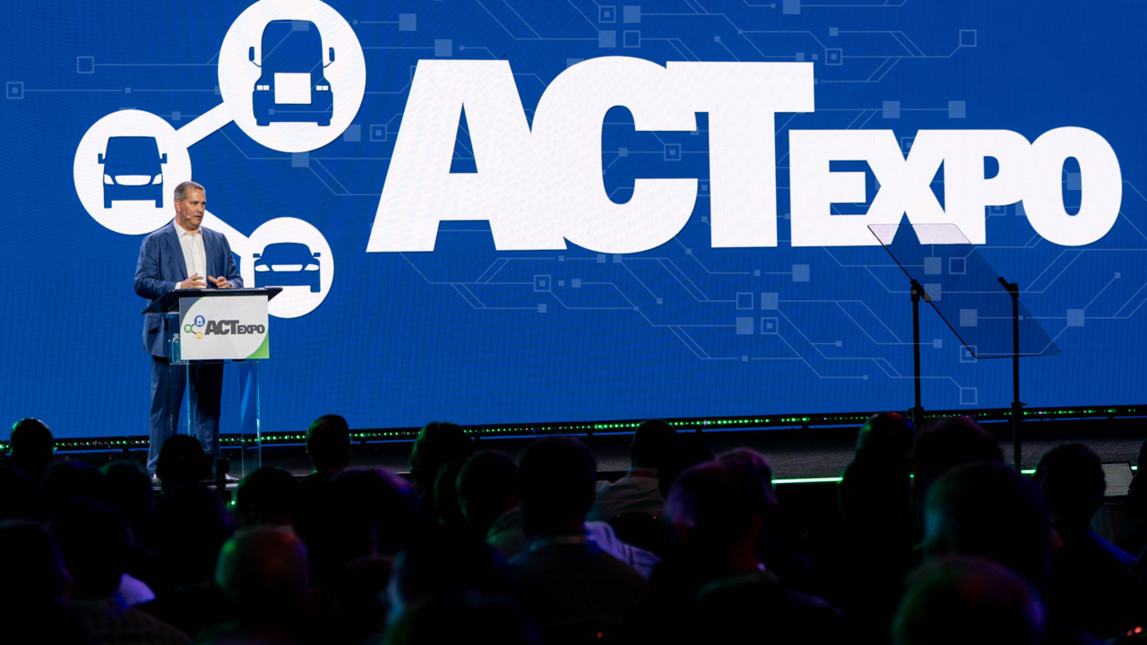A keynote speaker in a blue suit delivers an address from the podium on the main stage at ACT Expo, with the large ACT Expo logo, connected truck icons, and a digital circuit-themed blue backdrop behind him, as an engaged audience looks on in the foreground.