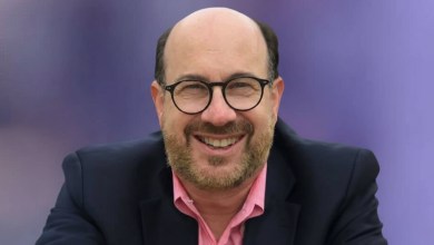 Photo of a bald man with dark glasses and dark hair on the side, wearing a pink shirt and navy blazer.