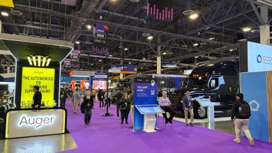 Manifest 2026 exhibition floor in Las Vegas featuring the Auger autonomous supply chain technology booth with its bright yellow signage and a large black semi-truck display amid attendees on the purple-carpeted hall