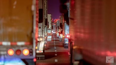 Line of traditional semi-trucks queued in nighttime highway traffic, red taillights glowing against a dark purple sky