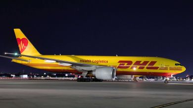 A mustard-yellow DHL plane with red lettering and "Health Logistics" banner is parked at night on an airfield, as seen from the side.