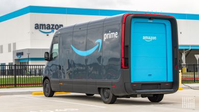 A two-tone blue Amazon Prime delivery van is parked in front of an Amazon distribution center.