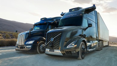 Aurora autonomous trucks: A Volvo and Peterbilt self-driving semi-truck with rooftop sensors parked side by side on a desert road at sunset.