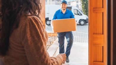 A courier with a hat and blue shirt delivered a box as seen through the front door.