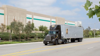 A brown UPS tractor pulls a trailer along a road next to a warehouse.