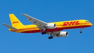 A mustard-yellow plane with red DHL lettering comes in for a landing as seen from below.