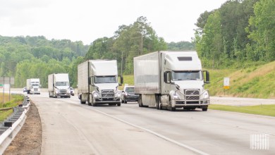 White semi-trucks traveling in convoy on a multi-lane highway with green hills and trees in the background.