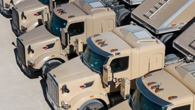 Beige Kodiak autonomous semi-trucks equipped with roof-mounted sensor pods and antennas, parked closely together in a sunny outdoor lot.