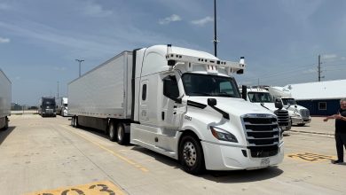 Bot Auto's white autonomous Freightliner truck with roof-mounted sensors, parked in a sunny Texas yard with a person gesturing on the right.