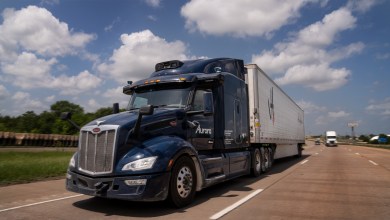 Blue Peterbilt Aurora autonomous truck pulling white Hirschbach refrigerated trailer on sunny highway