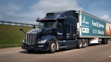 Dark blue Aurora autonomous semi-truck with McLane Food & Service trailer driving on a sunny Texas highway