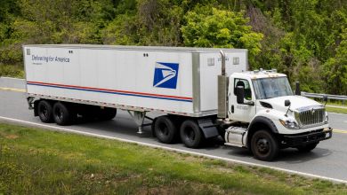 A U.S. Postal Service tractor-trailer truck on a road with green grass and trees around.