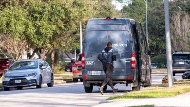 An Amazon delivery driver opens the door off the van on a neighborhood street with cars parked along the curb nearby.