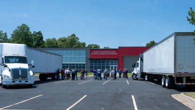 Two white semi-trucks with trailers donated by FedEx Freight to UA Northark, parked outside the Center for Robotics and Manufacturing Innovation building as a group gathers for the CDL training program relaunch.