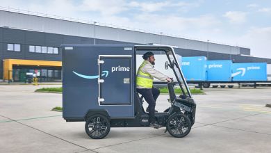 A driver operates a dark-blue Amazon e-cargo bike with an Amazon warehouse and light-blue trucks in the background.
