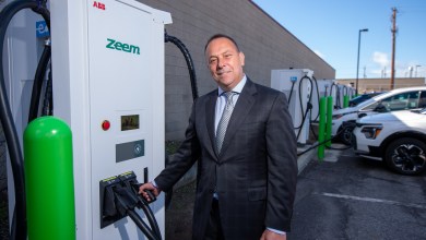 Zeem Solutions CEO Paul Gioupis stands next to a Zeem-branded ABB fast charger at a commercial electric vehicle charging depot with multiple chargers and fleet vehicles in the background.