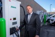 Zeem Solutions CEO Paul Gioupis stands next to a Zeem-branded ABB fast charger at a commercial electric vehicle charging depot with multiple chargers and fleet vehicles in the background.